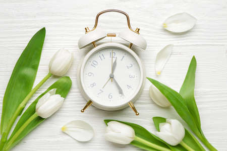 Alarm clock and flowers on white wooden background. Spring time