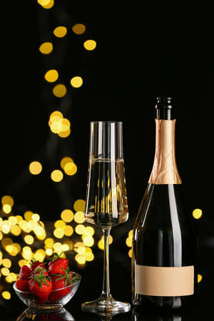 Glass and bottle of tasty champagne with strawberry on dark background with blurred lightsの写真素材