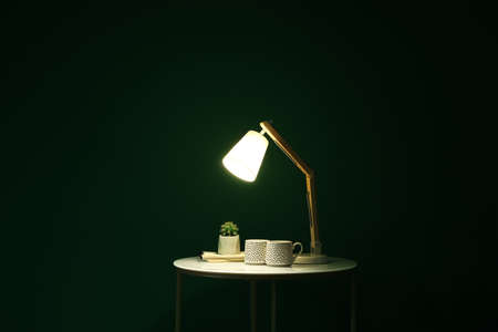 Glowing lamp, cups and houseplant on table in dark roomの写真素材