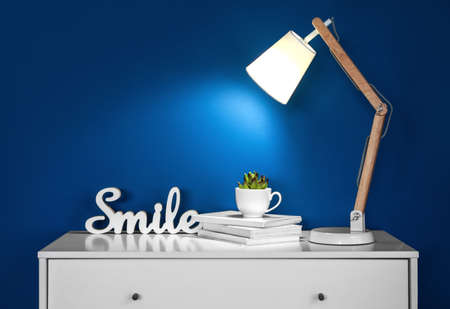 Chest of drawers with glowing lamp, books and houseplant in roomの写真素材