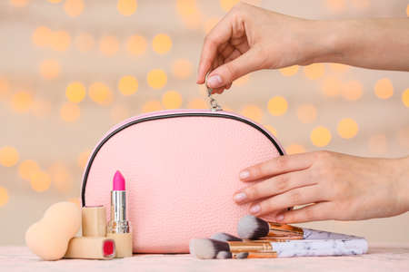 Female hands and bag with decorative cosmetics on color background with blurred lights