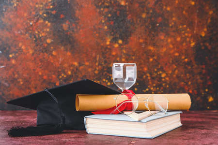 Graduation hat, book, hourglass and diploma on tableの写真素材