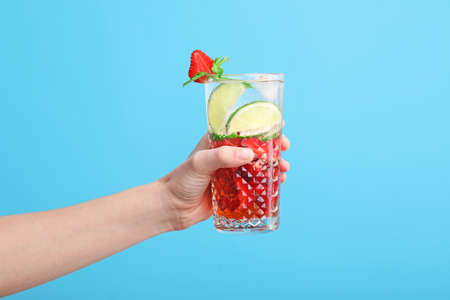 Hand with glass of fresh strawberry lemonade on color background