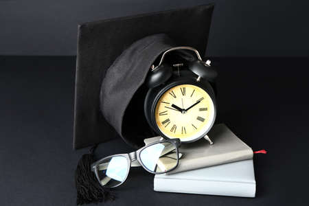 Graduation hat, alarm clock, eyeglasses and books on dark background