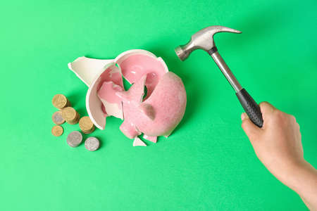 Hand with hammer and broken piggy bank on color backgroundの写真素材