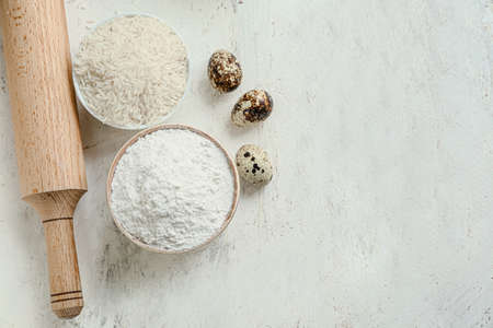 Bowls with rice, flour, eggs and rolling pin on white table