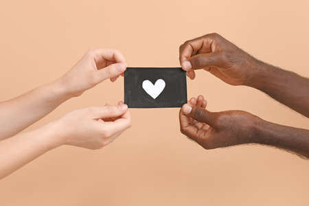 Hands of Caucasian woman and African-American man holding paper sheet with drawn heart on color background. Racism conceptの写真素材