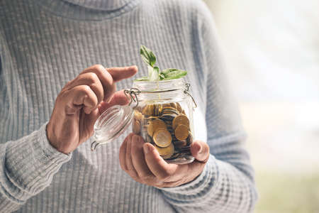 Senior woman with savings and young plant in jar near window at home. Concept of pensionの写真素材