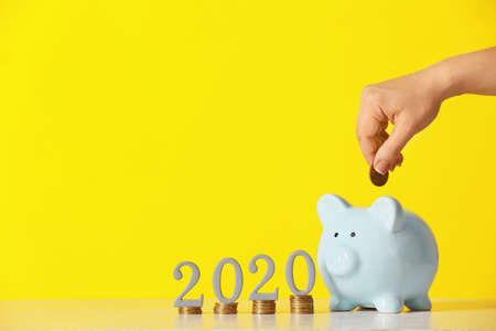 Female hand putting coin into piggy bank and figure 2020 on color background. Concept of pension