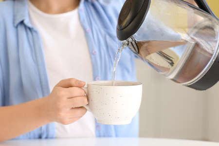 Young woman pouring boiled water from electric kettle into cup in kitchen, closeup
