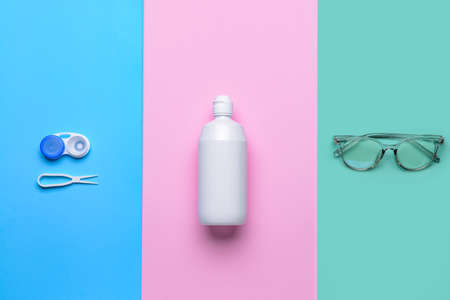 Container with contact lenses, solution tweezers and eyeglasses on color background
