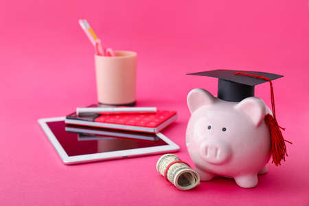 Piggy bank with graduation hat, money, tablet and stationery on color backgroundの写真素材