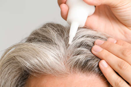Mature man using serum for hair loss treatment on gray background, closeup