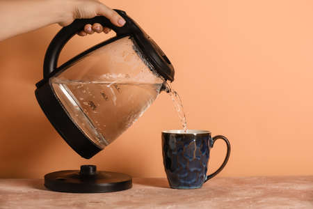 Woman pouring hot boiled water from electric kettle into cup on table