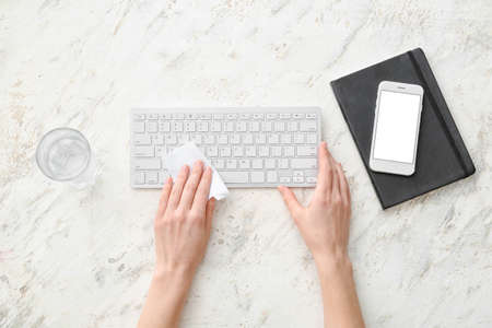 Woman cleaning computer keyboard, mobile phone and glass of water on light backgroundの写真素材