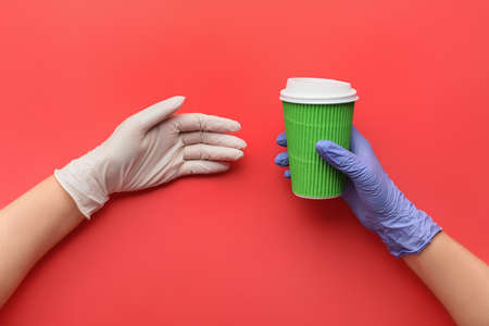 Hands in protective gloves and with cup of coffee on color background