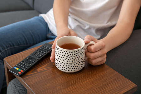 Woman with cup of tea watching TV on sofa at home