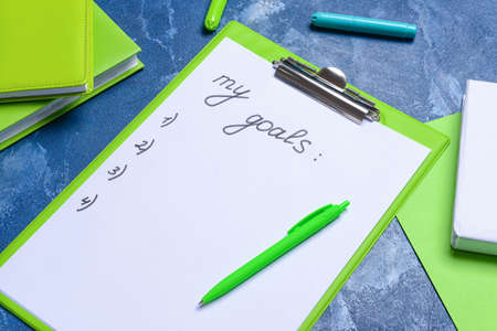 Clipboard with text MY GOALS and pen on color background