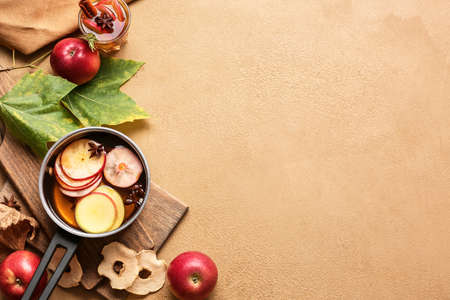 Tasty drink with spices and apple slices in cooking pot on color background