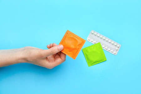 Female hand with wrapped condoms and birth control pills on color backgroundの写真素材