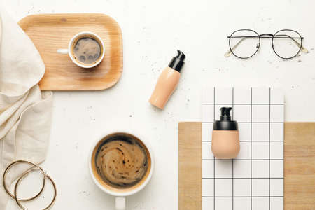 Bottles with makeup foundation and cups of coffee on light background