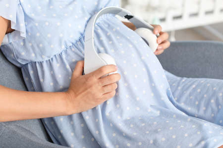 Beautiful pregnant woman putting headphones on her belly at home, closeup
