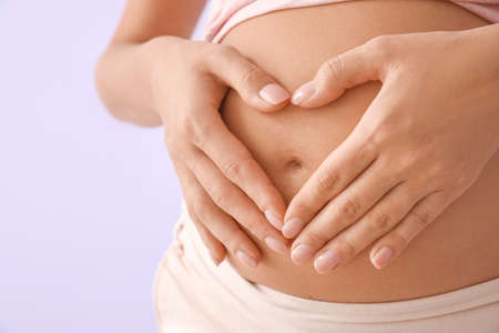 Beautiful pregnant woman making heart with her hands on color background, closeup