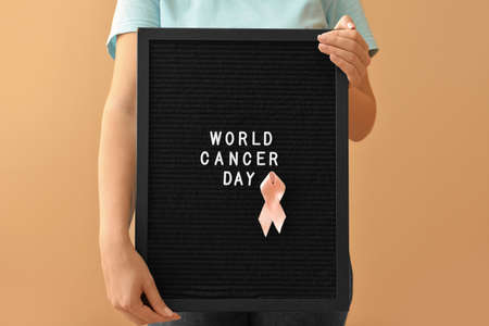 Woman holding board with text WORLD CANCER DAY and pink ribbon on color backgroundの写真素材