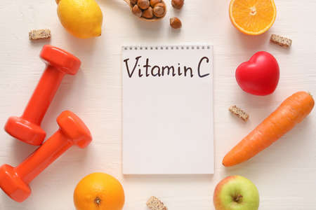 Healthy products, dumbbells and notebook with text VITAMIN C on light wooden background