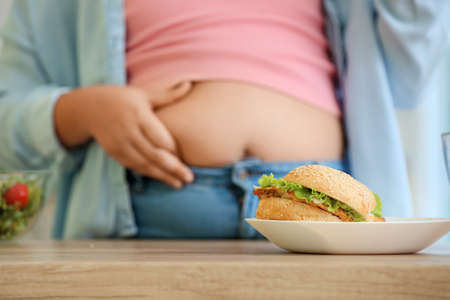 Plate with unhealthy burger on table near overweight girl