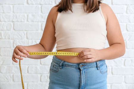 Overweight girl measuring her waist on white brick background