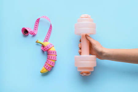 Female hand, dumbbell, banana and measuring tape on color background. Weight loss concept