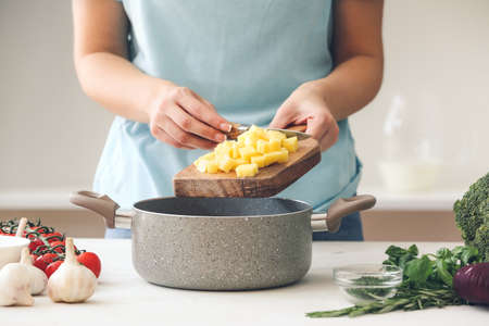 Woman putting cut potato into cooking pot on table in kitchenの写真素材