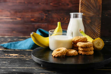 Board with tasty cookies, bananas and glass of milk on dark wooden backgroundの写真素材