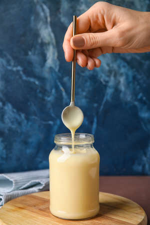 Female hand, spoon and jar with sweet condensed milk on color backgroundの写真素材