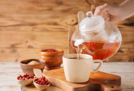 Woman pouring goji tea from teapot into cup on wooden backgroundの写真素材