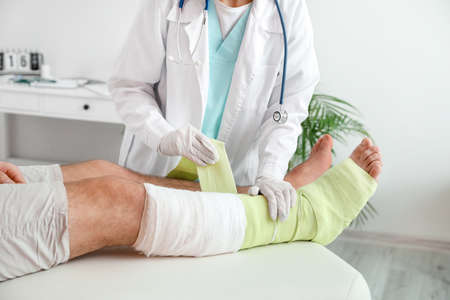 Doctor applying bandage on broken leg of young man in clinicの写真素材