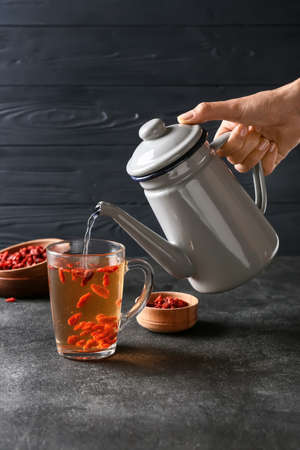 Woman pouring tasty tea with goji berries from teapot into glass cup on dark backgroundの写真素材