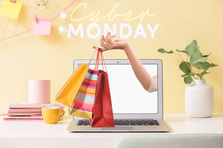 Woman sticking hand with shopping bags out of laptop screen on table in office. Cyber Monday Saleの写真素材