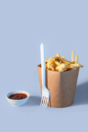 Box with tasty french fries, fork and sauce on color background