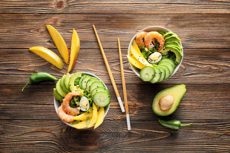 Bowls of fresh salad with mango, shrimps and vegetables on wooden background