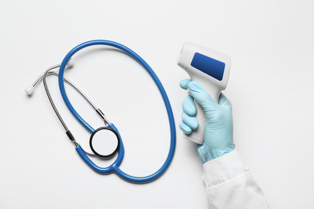Hand of doctor with infrared thermometer and stethoscope on light background