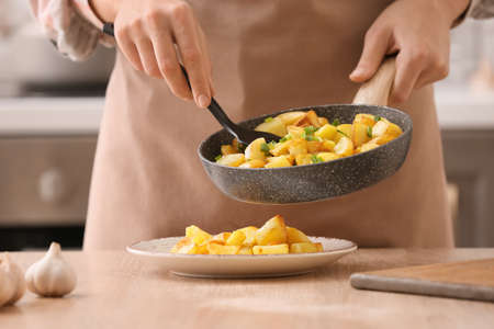 Woman putting tasty potatoes from frying pan into plate in kitchenの写真素材