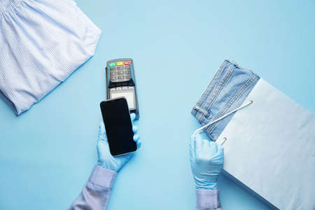 Female hands with mobile phone, payment terminal and clothes on color background