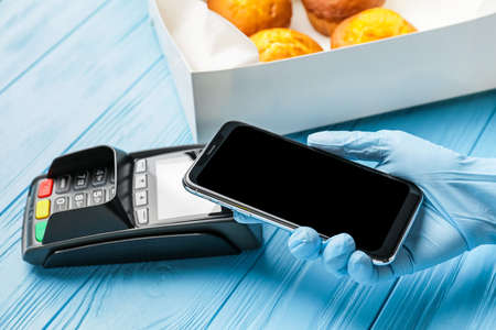 Female hand with mobile phone, payment terminal and tasty cupcakes in box on color wooden background