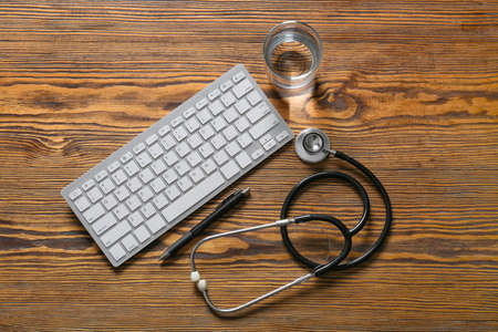 Modern stethoscope, computer keyboard, glass of water and pen on wooden backgroundの写真素材