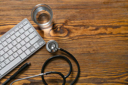 Modern stethoscope, computer keyboard, glass of water and pen on wooden backgroundの写真素材
