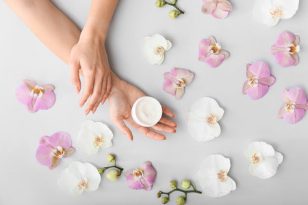 Female hands with cream and beautiful flowers on light backgroundの写真素材