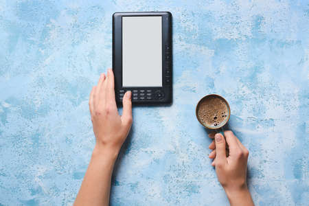 Female hands with e-reader and cup of coffee on color backgroundの写真素材