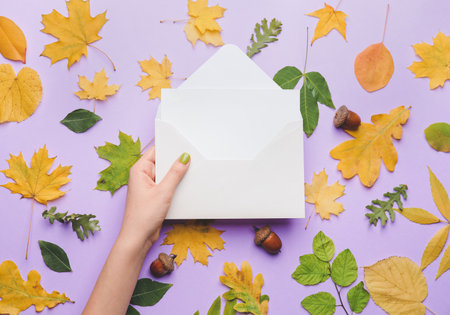 Female hand with envelope, blank card and autumn leaves on color background